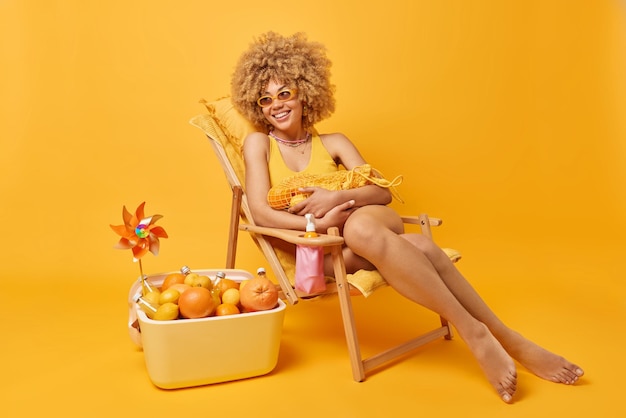 嬉しい女性は柑橘系の果物が入ったネットバッグを持って幸せそうに見えます水着のサングラスに身を包んだデッキチェアに座って黄色い壁のポータブルクーラーの近くで隔離された良い夏の日完璧な休暇を楽しんでいます