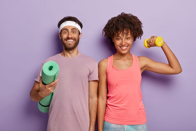 Free photo glad woman and man of different nationality, stand shoulder to shoulder, carry karemat, raise dumbbell