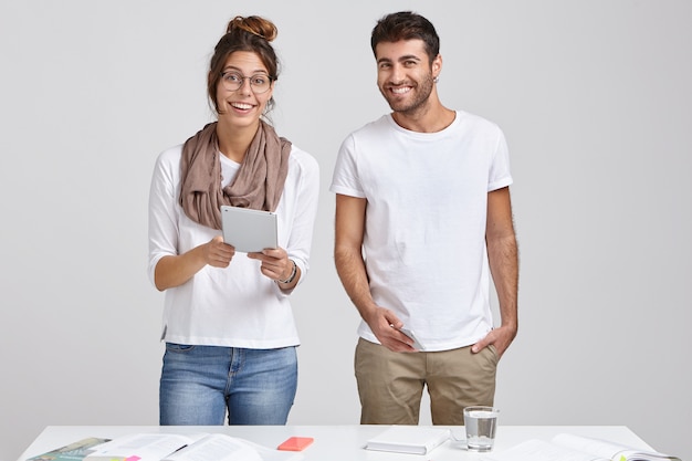 Glad woman and man students future architects, use modern tecnologies for work, stand at table with necessary papers, have happy expressions, ready to start preparation for seminar