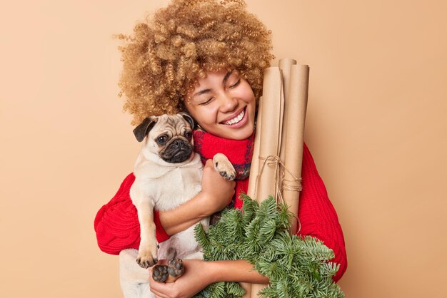 Радостная женщина счастливо улыбается, с любовью обнимает собаку, собирающуюся отпраздновать Новый год или Рождество, держит еловый венок и свернутую бумагу, изолированные на бежевом фоне. Счастливый владелец домашнего животного держит предметы для украшения
