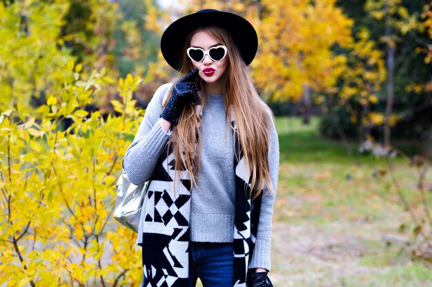 嬉しい女性は晴れた秋の日にエレガントなコートとサングラスでポーズをとる黒い帽子を着ています。黄色の木のある公園を歩いてトレンディな灰色のセーターで熱狂的な女性モデルの屋外のポートレート。