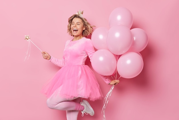 Glad woman wears festive dress has hairstyle holds magic wand and bunch of balloons has upbeat mood celebrates special occasion looks happily away isolated over pink background. Partying concept
