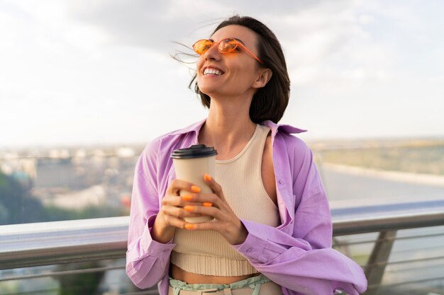 スタイリッシュな夏の衣装で短い髪の嬉しい女性は、モダンな橋でコーヒーを飲みます