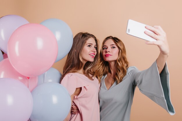 Glad woman with trendy wavy hairstyle using phone for selfie with friend