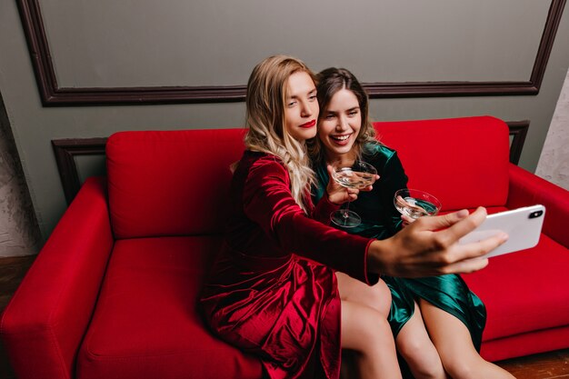 Glad women drinking wine on couch. Indoor portrait of winsome girls celebrating something at home.