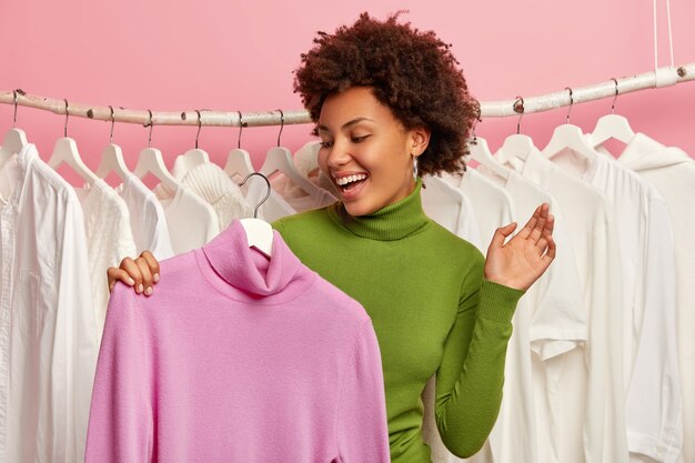 Glad young Afro woman happy to find jumper, holds purple solid turtleneck on hanger, chooses outfit for party, does shopping in boutique, keeps palm raised