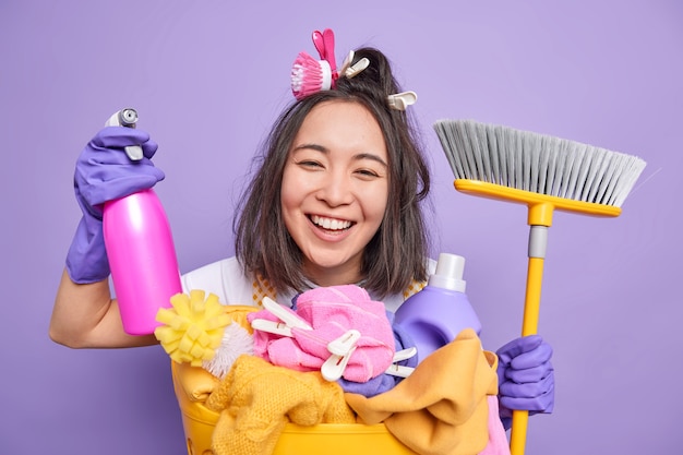 Glad young Asian housewife smiles broadly helps about house wears rubber gloves holds cleaning detergent and broom for sweeping floor poses near laundry basket isolated over purple background