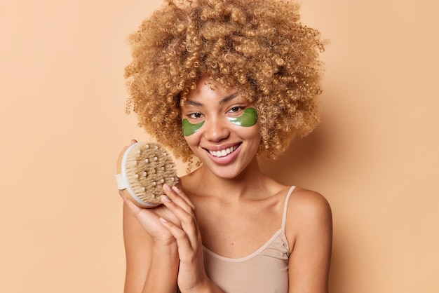 Free photo glad young curly haired female model wears casual t shirt holds body brush undergoes skin care treatments smiles pleasantly poses indoor against beige background spa care and cosmetology concept