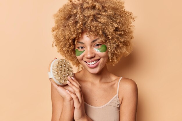 Glad young curly haired female model wears casual t shirt holds body brush undergoes skin care treatments smiles pleasantly poses indoor against beige background Spa care and cosmetology concept
