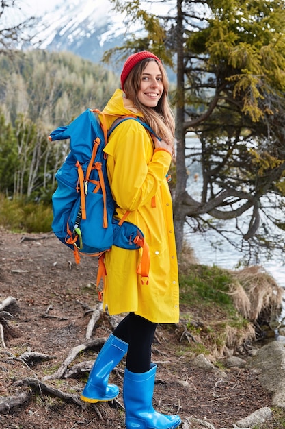 Glad young female model with backpack wears red headgear, yellow raincoat and rubber blue boots