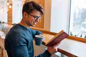 Free photo glad young man resting at cafe, reading interesting book, drinking tea.