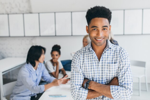 他の従業員と彼のオフィスで腕を組んでポーズをとるアフリカの髪型のうれしい若い男。職場での会議中に笑顔の青いシャツの男性マネージャー。