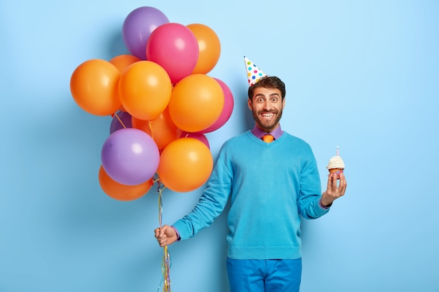 Glad young man with bristle, holds delicious small muffin, bunch of colored balloons