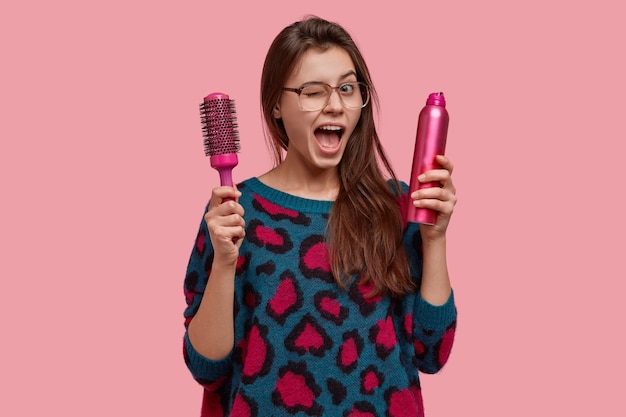 Glad young woman blinks eyes, keeps jaw dropped, carries hairbrush and spray, ready to make nice hairstyle for you Free Photo