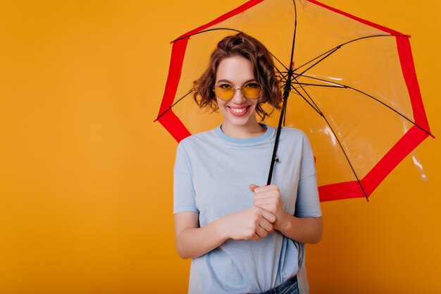 Glad young woman in blue t-shirt holding stylish parasol. Indoor portrait of pleased curly girl in sunglasses posing with smile under umbrella.