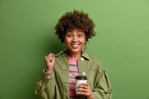 기쁜 젊은 여자 clenches 주먹 긍정적 인 뉴스 미소를 축하 행복하게 음료 테이크 아웃 커피 녹색 벽 위에 고립 된 세련된 옷을 입고