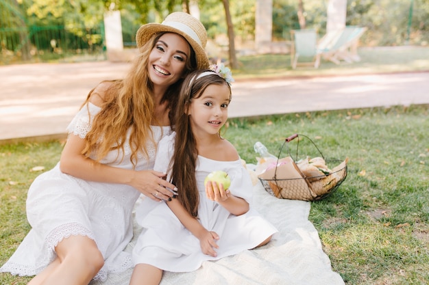 Glad young woman in elegant attire gently embracing girl, eating green apple with appetite. Outdoor portrait of happy family having lunch in park and joking.