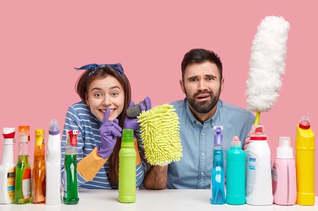 Glad young woman makes silence gesture, asks not spread rumors, sits near frustrated bearded husband, surrounded with detergents