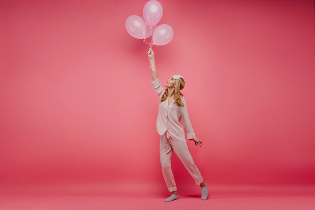 Felice giovane donna in pigiama di seta ballando con palloncini festa. foto a figura intera della spettacolare ragazza di compleanno in costume da notte divertente in posa sul muro rosa