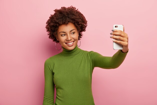 Glad young woman takes picture of herself on her smartphone, smiles pleasantly, dressed in green turtleneck jumper, has Afro hairstyle, models against pink wall. Time for selfie.