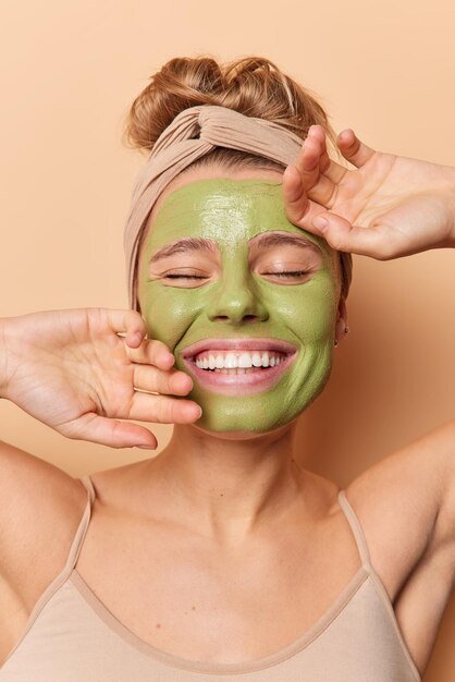 嬉しい若い女性が顔に優しく触れるスキンケアのための緑の美容マスクを適用し、目を閉じたままにしますヘッドバンドを着用しますTシャツはベージュの背景の上に分離されたスキンケア治療を受けます。ウェルネスコンセプト