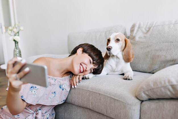 Glad young woman in trendy dress sitting beside sofa and making selfie with her puppy