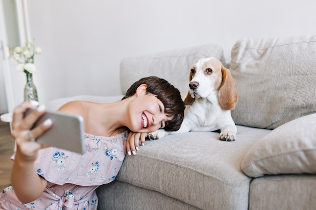 Glad young woman in trendy dress sitting beside sofa and making selfie with her puppy