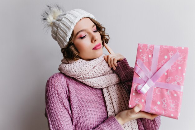 기쁜 젊은 여자는 새 해 선물을 들고 니트 흰 모자를 착용합니다. 귀여운 리본으로 장식 된 분홍색 선물 상자와 함께 포즈 멋진 여성 모델.