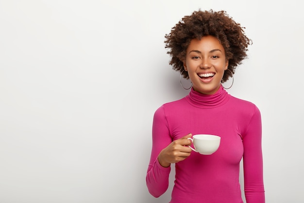 Free photo glad young woman wears pink poloneck, holds mug with coffee, enjoys spare time for live communication with friend