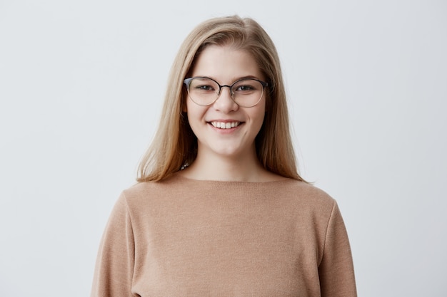 Felice giovane donna con i capelli lisci biondi e gli occhiali, con un sorriso gentile essendo felice di incontrare il suo ragazzo. femmina dello studente che sorride nella macchina fotografica che si rallegra il suo successo all'università.