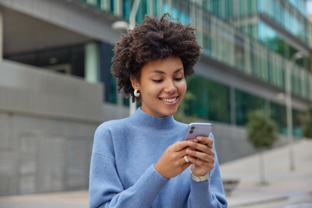 巻き毛のふさふさした髪のうれしい若い女性は、現代の携帯電話のダウンロードを保持しています驚くべきアプリケーションサーフィンソーシャルネットワークはカジュアルな青いジャンパーを着ています