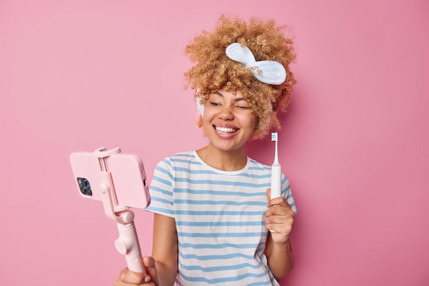 Glad young woman with curly hair wears headband and casual striped t shirt records video blog holds smartphone on selfie stick gives recommendations how to care about teeth holds toothbrush