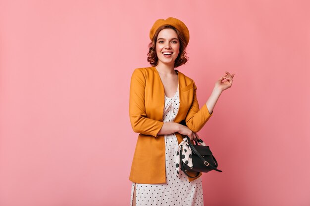 Glad young woman with handbag looking at camera. Studio shot of amazing french girl in beret isolated on pink background.