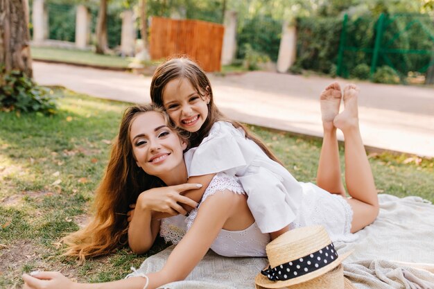 Glad young woman with sparkle make-up chilling on blanket with legs up and smiling. Laughing tanned girl lying on mother's back