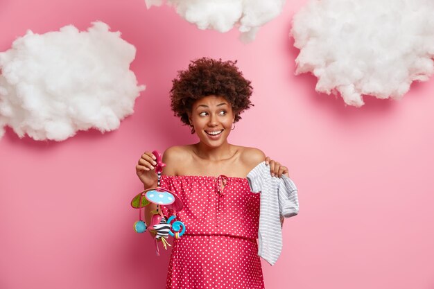 Gladsome ethnic woman poses with baby bodysuit and mobile, will become mother soon, looks happily aside, has big belly, wears polka dot dress, isolated on pink wall, white clouds overhead