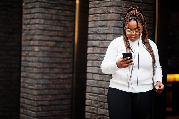 Гламурная африканская американка в белом свитере с высоким воротом позирует на улице с мобильным телефоном