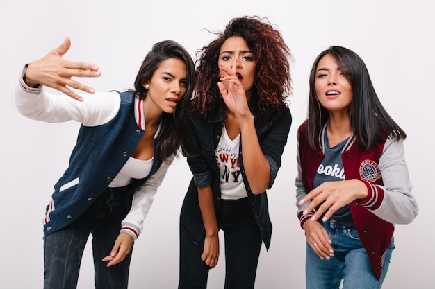 Glamorous african girl covers mouth with hand posing with unhappy face expression. Pretty asian lady with straight hair having fun with best friends in stylish attires.