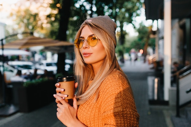 Glamorous blonde woman in round sunglasses carrying cup of coffee and looking away in front of shady street