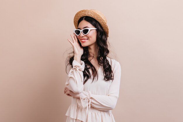 Glamorous brunette woman laughing on beige background. Studio shot of blissful young woman in straw hat.