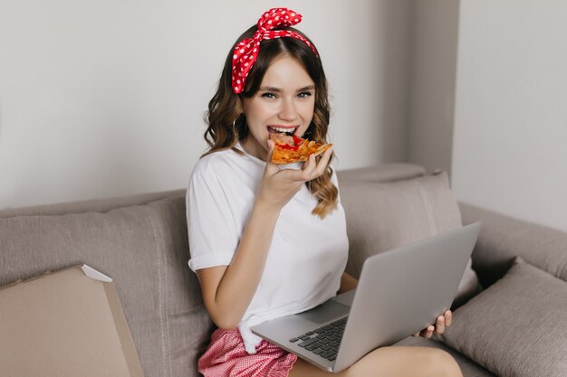 Гламурная кудрявая девушка с удовольствием ест пиццу и пользуется компьютером. Привлекательная женщина, проводящая выходные дома с ноутбуком и фаст-фудом.