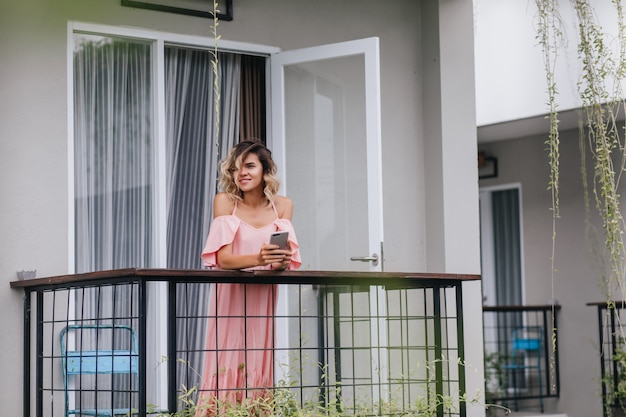 Free photo glamorous curly lady standing at balcony with smartphone. charming caucasian girl looking at city from terrace.