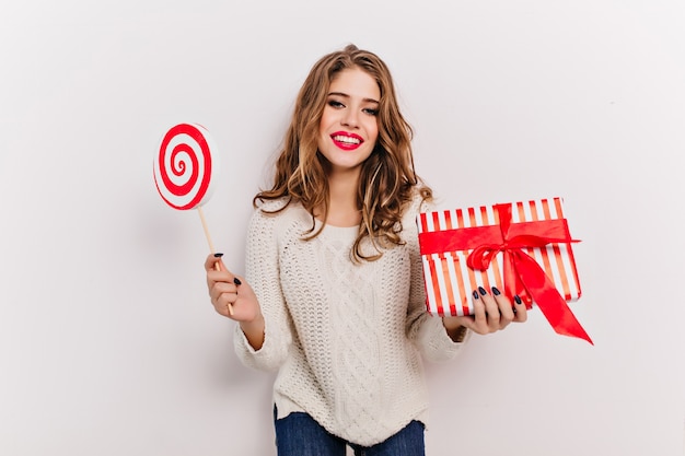 Glamorous european woman in stylish sweater holding new year present and laughing. Indoor portrait of curly girl posing with lollipop and box decorated with ribbon.