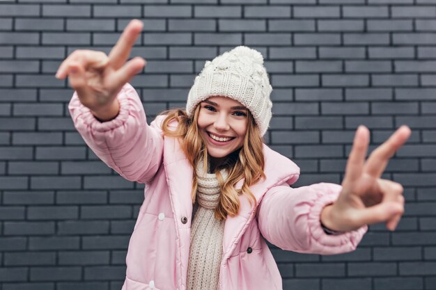 Гламурная женская модель в розовой зимней куртке позирует со знаком мира. Открытый выстрел смеясь над веселой блондинкой в вязаной шапке.