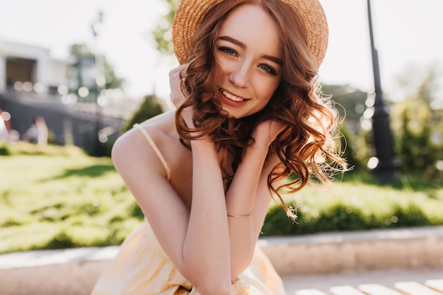 Glamorous female model with ginger hair posing with cute smile on nature. Outdoor photo of good-humoured girl in straw hat enjoying photoshoot in park.