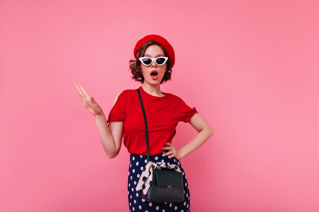 Glamorous french woman in red t-shirt posing. Indoor photo of brunette european girl in beret and sunglasses.