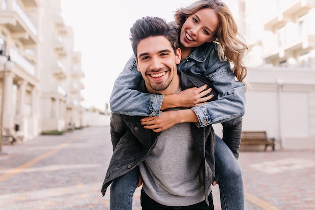 Glamorous girl sitting on boyfriend's back. Outdoor portrait of carefree caucasian couple posing in city