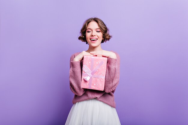 Glamorous girl with curly short hair posing with pink present box and laughing. Attractive female model with christmas gift isolated on purple wall and smiling.