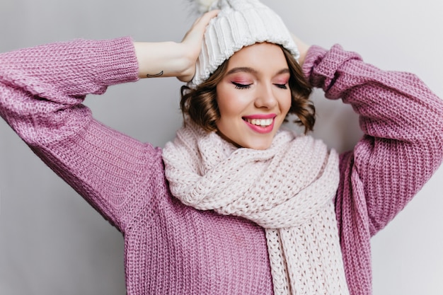 目を閉じてスカーフでポーズをとる短い散髪の魅力的な女の子。冬の服装で写真撮影を楽しんでいるニット帽のリラックスした白人女性。