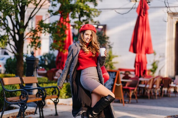 Glamorous lady in elegant knee high boots dancing in park with cup of coffee