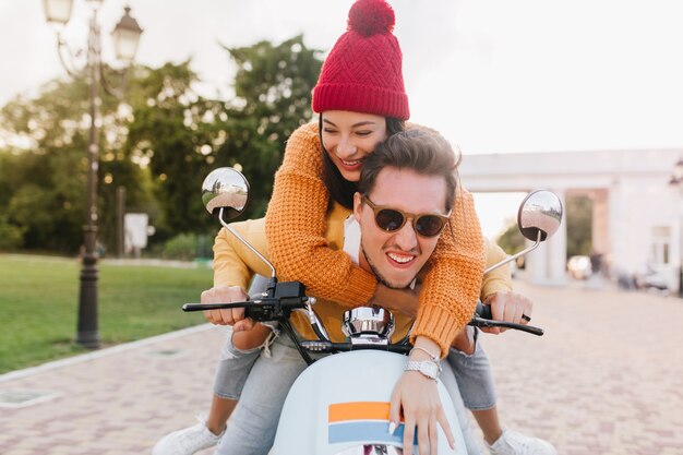 Glamorous woman in cute knitted hat looking away with smile, riding on scooter with boyfriend